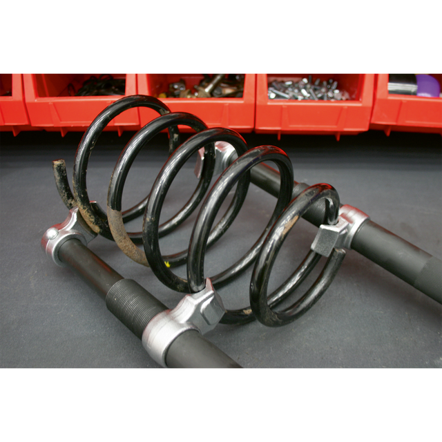 A close-up of the Sealey Coil Spring Compressor Set 2pc Heavy-Duty 2500kg/Pair - AK3845 on a workshop surface, with red storage bins containing various tools and parts in the background.