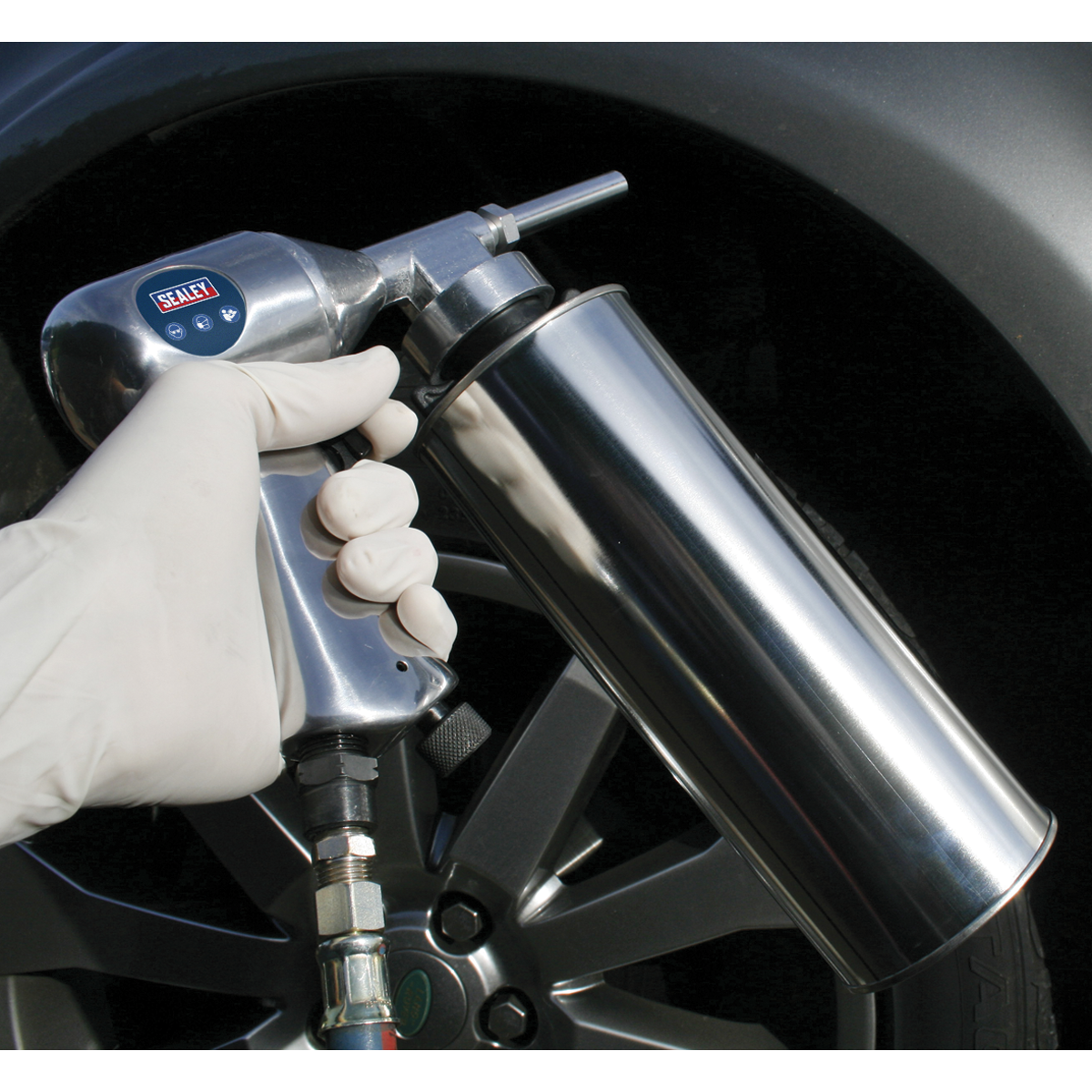 A gloved hand holds Sealey's Air Underbody Coating Gun - AK42, connected to a regulator set at 90psi, aimed at the tire of a vehicle.