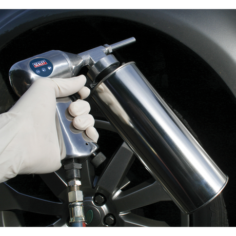 A gloved hand holds Sealey's Air Underbody Coating Gun - AK42, connected to a regulator set at 90psi, aimed at the tire of a vehicle.