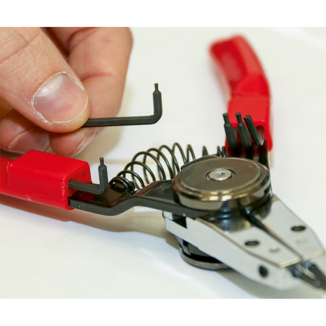 A hand adjusts the Sealey Circlip Pliers Set Internal/External Quick Change (AK8453), made from chrome vanadium steel and featuring red handles, using a small L-shaped hex key. This premier hand tool includes a spring mechanism and a circular component.