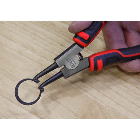 A hand holding the Sealey Circlip Pliers from the 180mm 4-piece set (AK8457) grips a circular metal ring on a wooden surface, showcasing the quality of Premier Hand Tools.