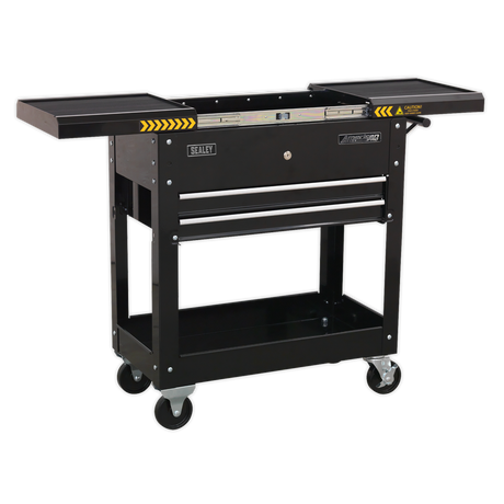 The Sealey Mobile Tool & Parts Trolley - Black - AP705MB features a single drawer, an open storage space beneath, and fold-out side trays. Constructed from heavy gauge steel, it comes with a lockable sliding top and heavy-duty ball-bearing slides. The trolley is equipped with four caster wheels and caution markings on the side trays.