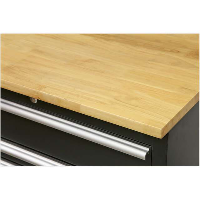 A close-up view of a Sealey Hardwood Worktop 1550mm - APMS07 with a smooth surface and the top of metal handles on black drawers beneath it, showcasing the details of Model No's APMS02 floor cabinets.