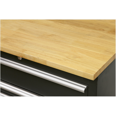 A close-up view of a Sealey Hardwood Worktop 1550mm - APMS07 with a smooth surface and the top of metal handles on black drawers beneath it, showcasing the details of Model No's APMS02 floor cabinets.