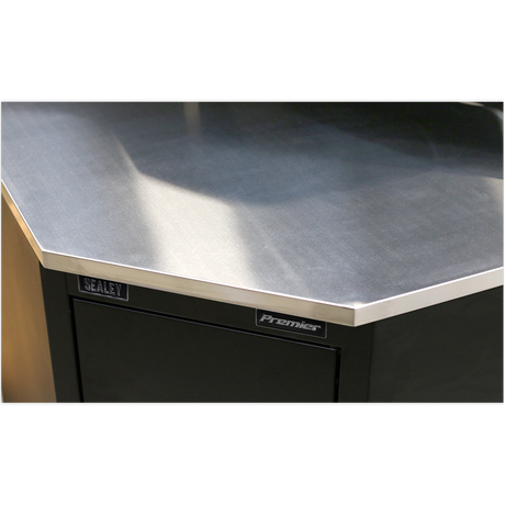 Close-up of a corner of a metallic workbench with "Sealey" label on a dark-colored cabinet, featuring a stainless steel worktop and Model No. APMS19 Stainless Steel Corner Worktop 930mm.