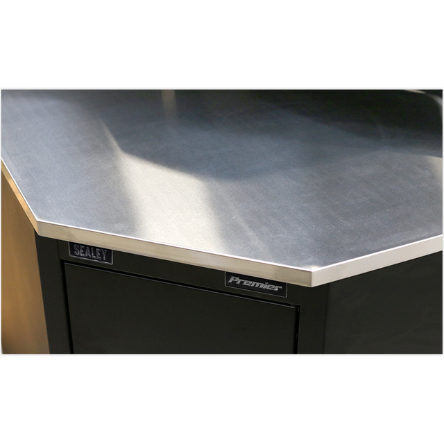 Close-up of a corner of a metallic workbench with "Sealey" label on a dark-colored cabinet, featuring a stainless steel worktop and Model No. APMS19 Stainless Steel Corner Worktop 930mm.