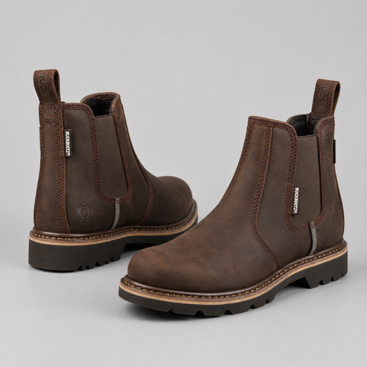 A pair of dark brown Chelsea boots with elastic side panels and pull tabs, shown from the front and side on a light gray background. The brand name 'KORSMUIR' is visible on the side tag.