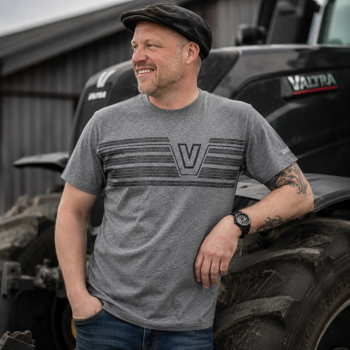 A man wearing a heather gray Valtra t-shirt with a dark gray striped graphic and 'V' logo, standing in front of a black Valtra tractor.