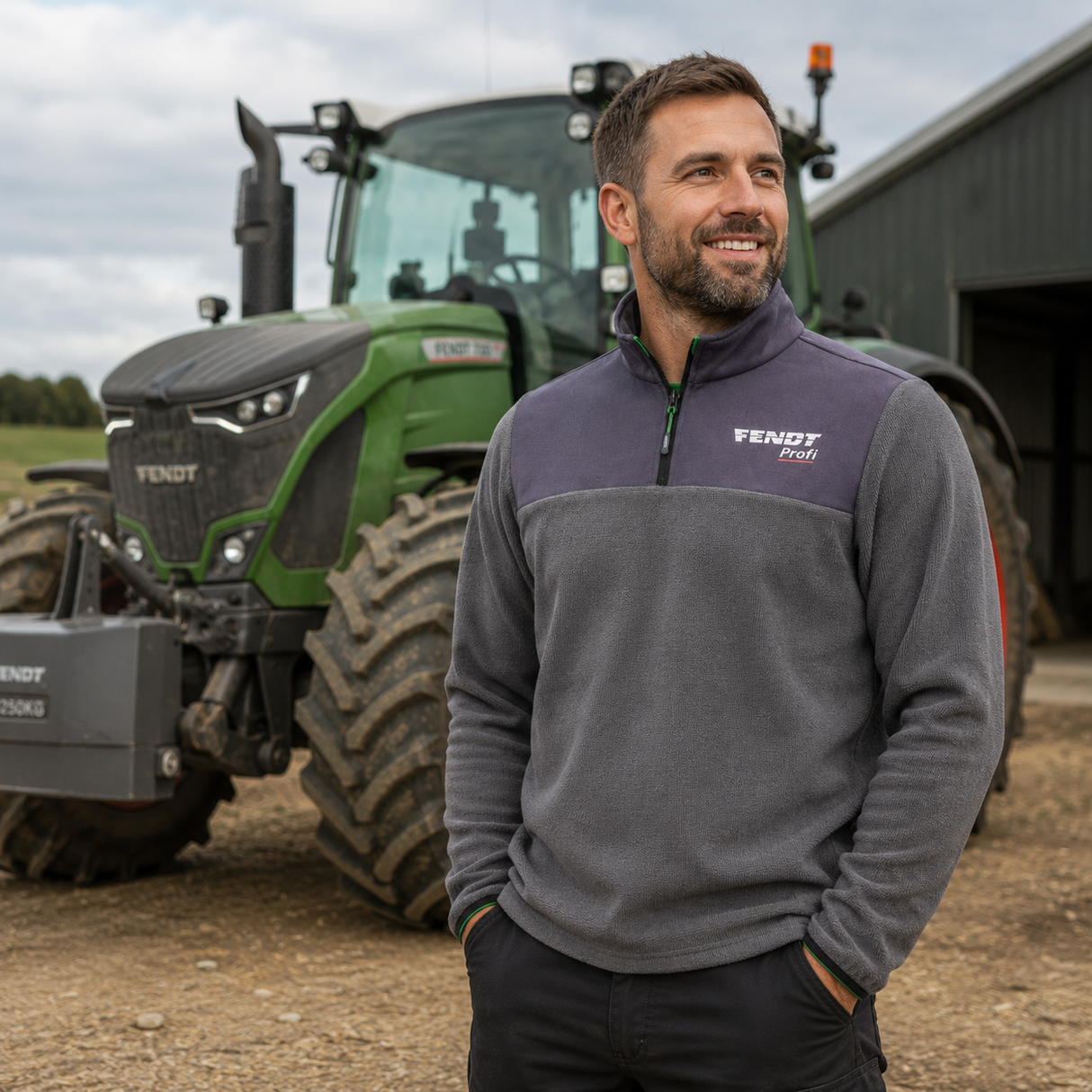 A smiling man stands outdoors in front of a green Vendy tractor and barn, wearing a gray and dark purple quarter-zip fleece pullover with a white Vendy Foli logo on the chest.