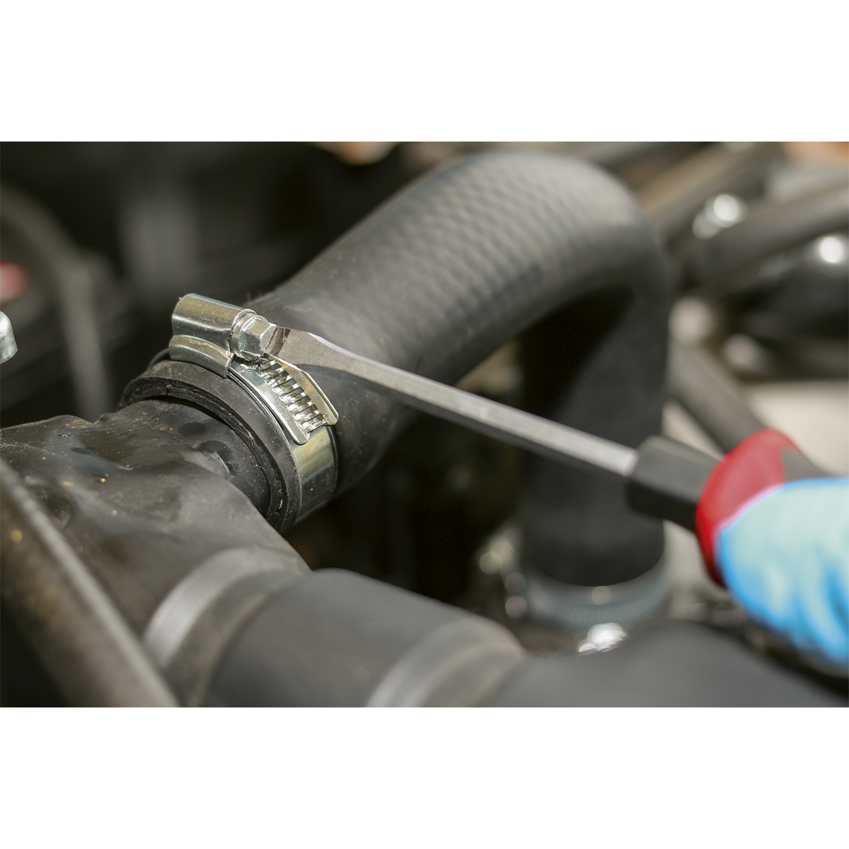 A person with a gloved hand is using a screwdriver to tighten a Sealey HI-GRIP® Hose Clip Zinc Plated Ø80-100mm (Pack of 10 - HCJ4X) on a black rubber hose in an automotive engine.