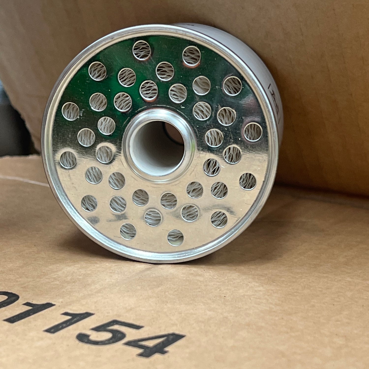 A cylindrical metal component with multiple holes on one end, resembling the Sparex Fuel Filter - Element - FF167A (Sparex Part No. S.109023), is carefully placed on a cardboard surface.