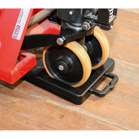 Close-up of the secure wheels on a red industrial jack, positioned on the Sealey Pallet Truck Stop - PTS1, featuring a non-slip durable rubber pad placed on a wooden floor.
