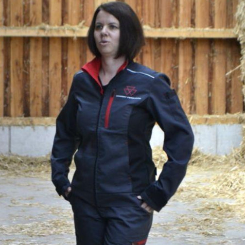 A person wearing the AGCO Massey Ferguson Women's Work Jacket (X993532214), featuring adjustable cuffs and reflective stripes, stands on a concrete floor in a barn with hay scattered in the background.