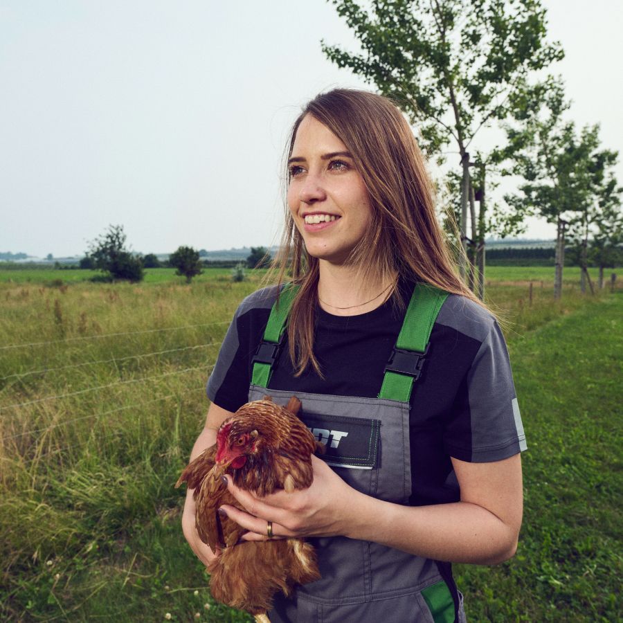 A woman standing outdoors in a grassy field wearing gray and green farm overalls over a black t-shirt, while holding a brown chicken.