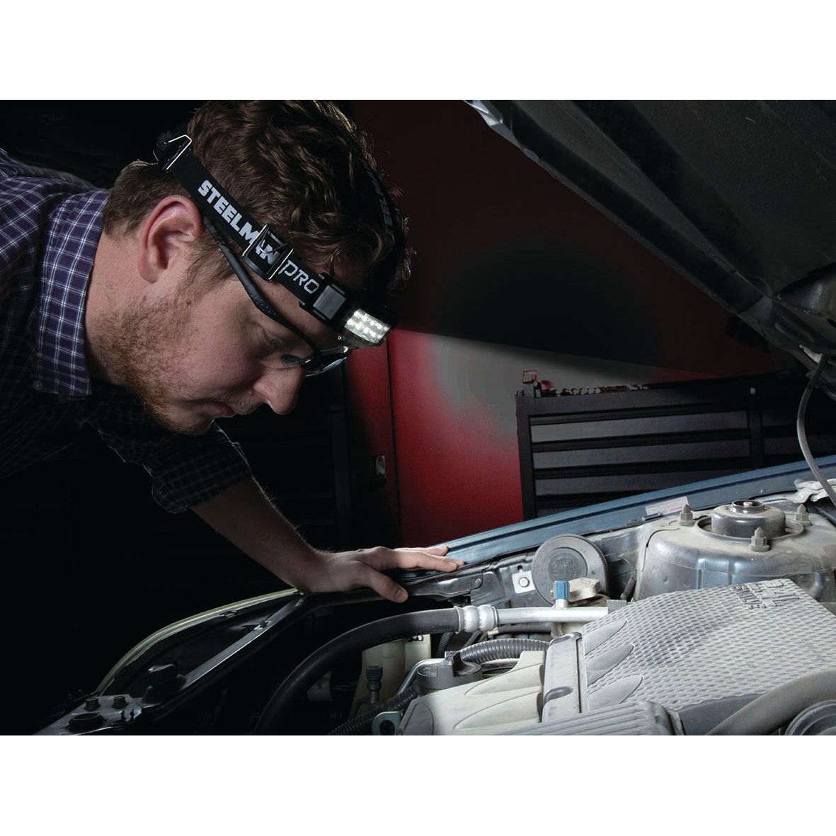 A person wearing the Slim rechargeable headlamp with movement detector - S.151756 from Sparex inspects a car engine in a dimly lit garage, ensuring the rechargeable battery is functioning properly.
