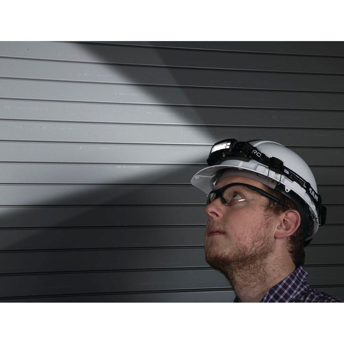 A man wearing a safety helmet and glasses looks upward with a Sparex Slim rechargeable headlamp with movement detector - S.151756, shining a focused beam on a metallic wall.