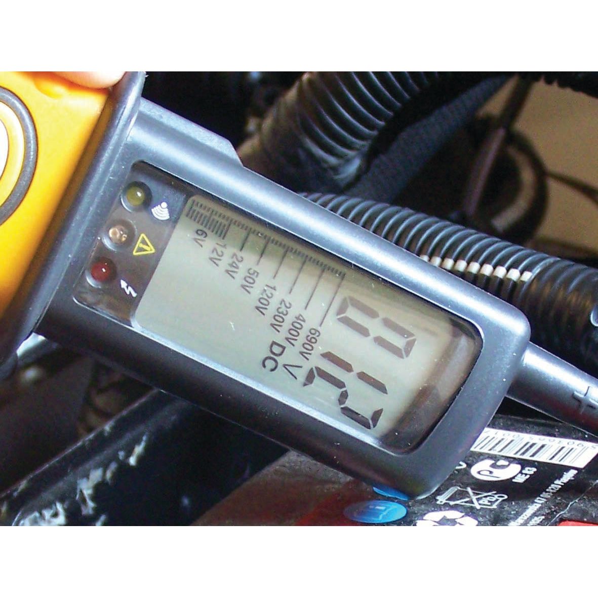 Close-up of an Electrical Voltage Tester - S.151758 from Sparex displaying a reading of 12.6 volts on a car battery. Various wires and vehicle components are visible in the background, highlighting the VAT voltage and continuity tester in action.