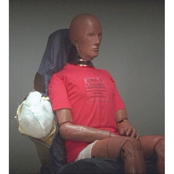 A crash test dummy in a red shirt, seated with arms on lap, is shown after an airbag deployment test. Next to the dummy's left side, you can see an inflated airbag protected by the Sparex Airbag Compatible Seat Cover - Car & Van - Universal Fit (Sparex Part No.S.71897).