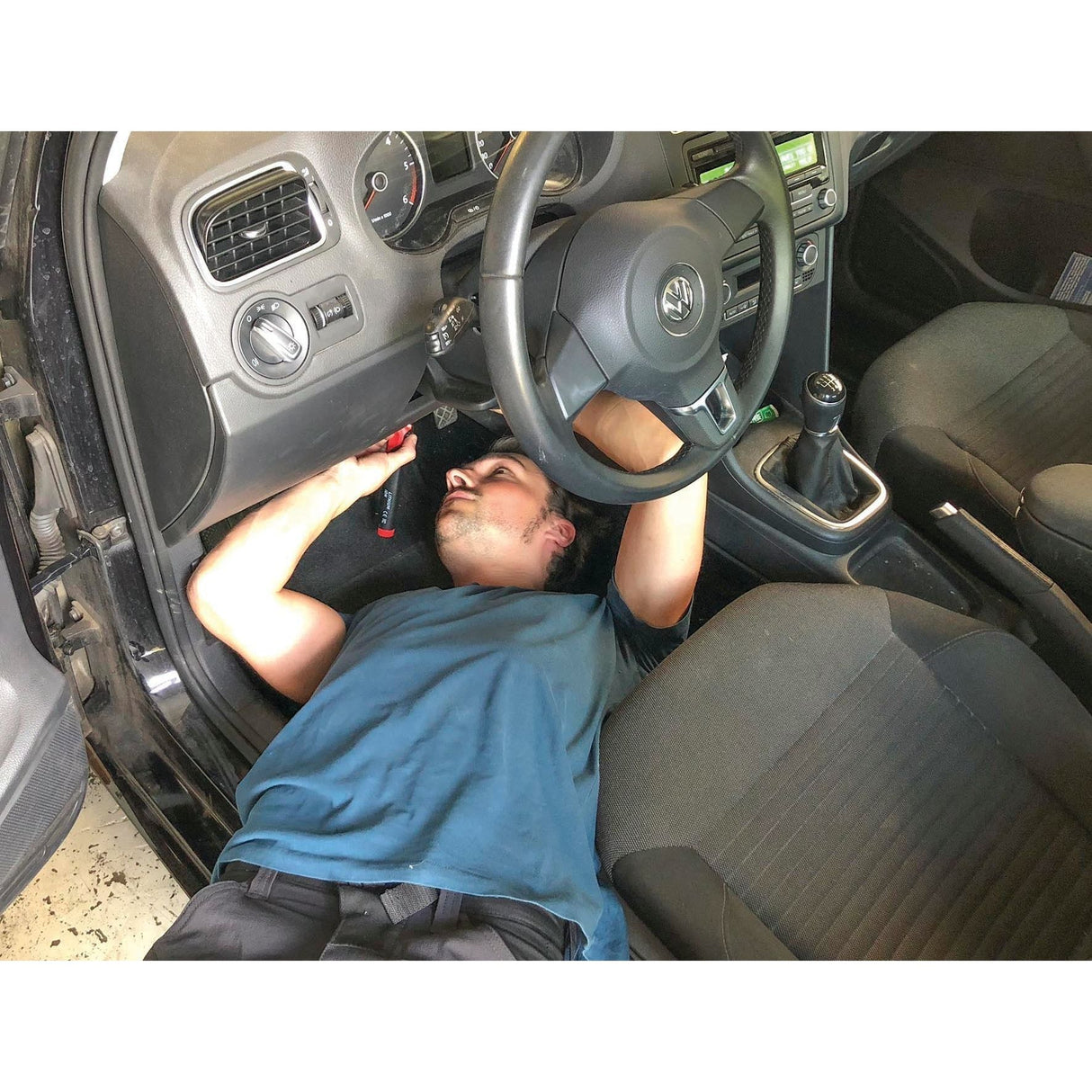 A person is lying on the floor of a car's driver-side with their upper body under the dashboard, seemingly working with tools, including a Sparex Cordless Soldering Iron - S.151757.