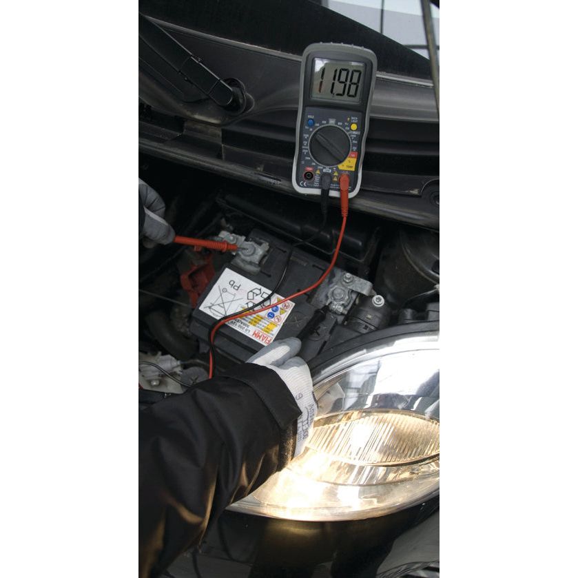 A Digital multimeter - S.151762 from Sparex displaying a Voltage DC of 11.98 is connected to a car battery under the hood. The engine bay is illuminated by a flashlight while a gloved hand holds the red probe against the battery terminal.