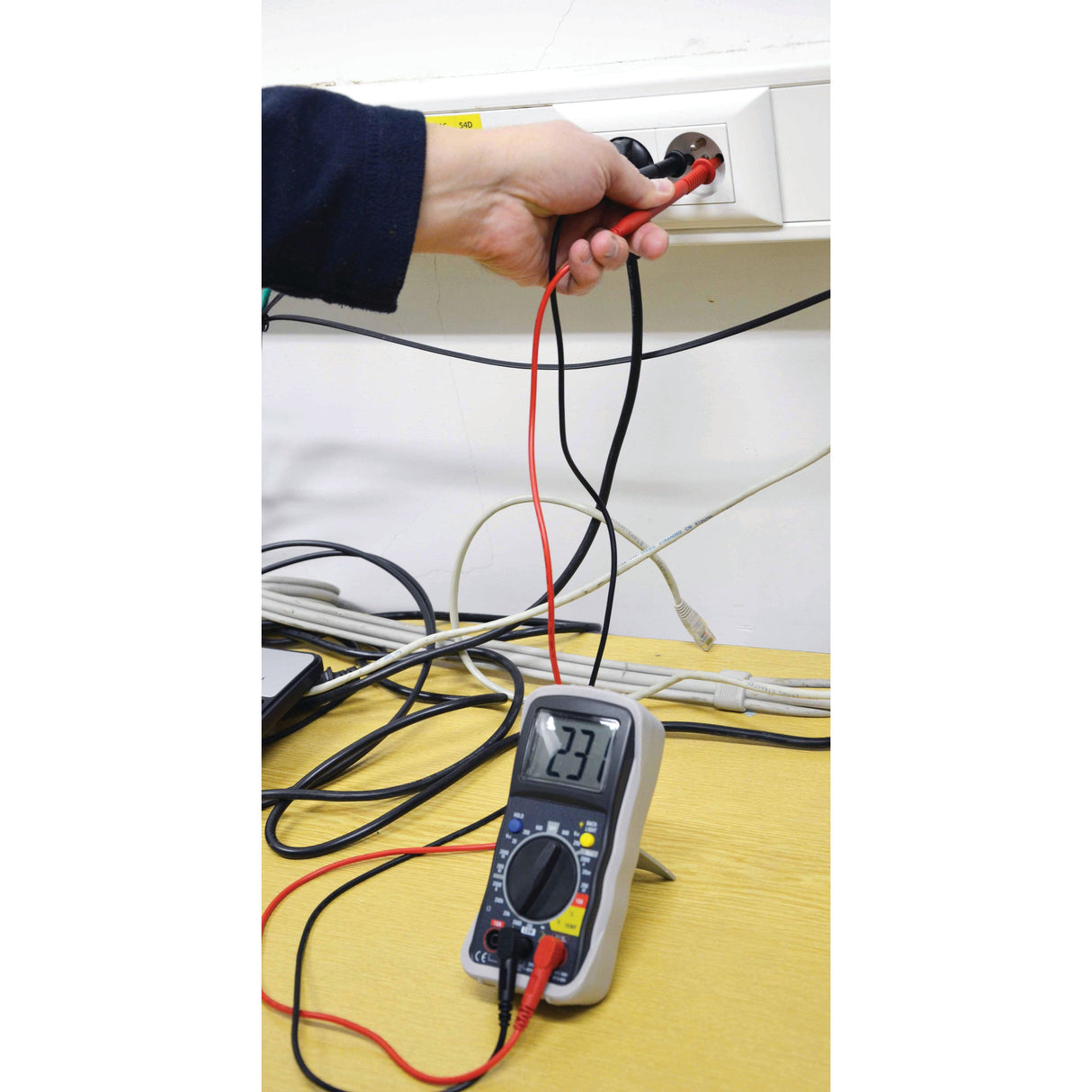 Using the Sparex Digital Multimeter - S.151762, a person measures the voltage of an electrical outlet, which displays 223 volts. The red and black probes are correctly connected to the outlet, ensuring accurate AC voltage measurement.