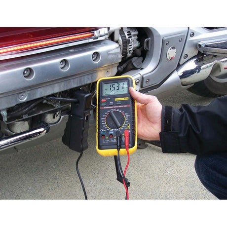 A person is holding a yellow Sparex Multimeter - S.151761 showing a reading of -15.37 near a vehicle's bumper, with black and red test leads connected to the vehicle for voltage measurement in automotive applications.