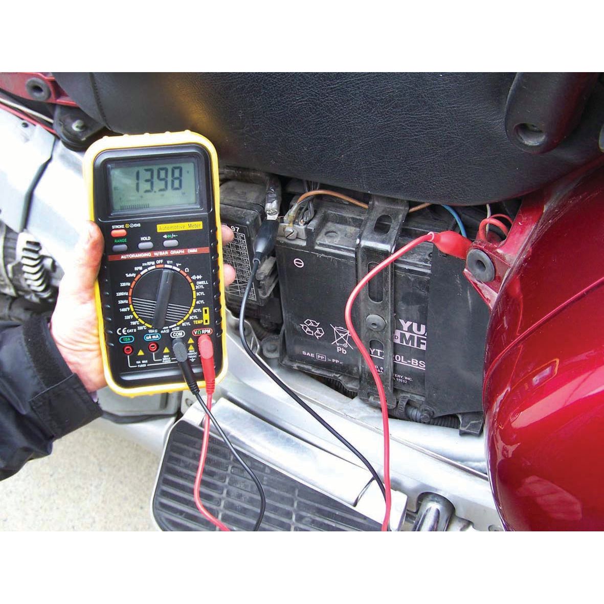 A person uses the Sparex Multimeter - S.151761 for voltage measurement on a motorcycle battery, showing a reading of 13.98 volts – an essential task in various automotive applications.