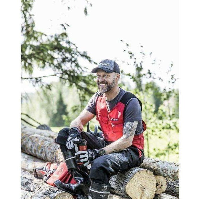 A man in a red vest and black boots sits on stacked logs outdoors, smiling, with a chainsaw and safety gear beside him. The text "Massey Tractor Parts" is visible in the bottom left corner. His AGCO Valtra Sleeveless Overalls - V4280530 feature kneepad pockets, perfect for a hard day's work.