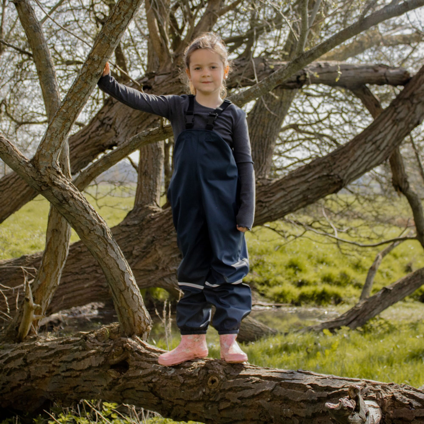 A young child dressed in Cottonmount's Fort Kids Splashflex PU Waterproof Bib n Brace Navy and pink boots stands on a large tree branch in an outdoor setting, holding onto one of the branches.