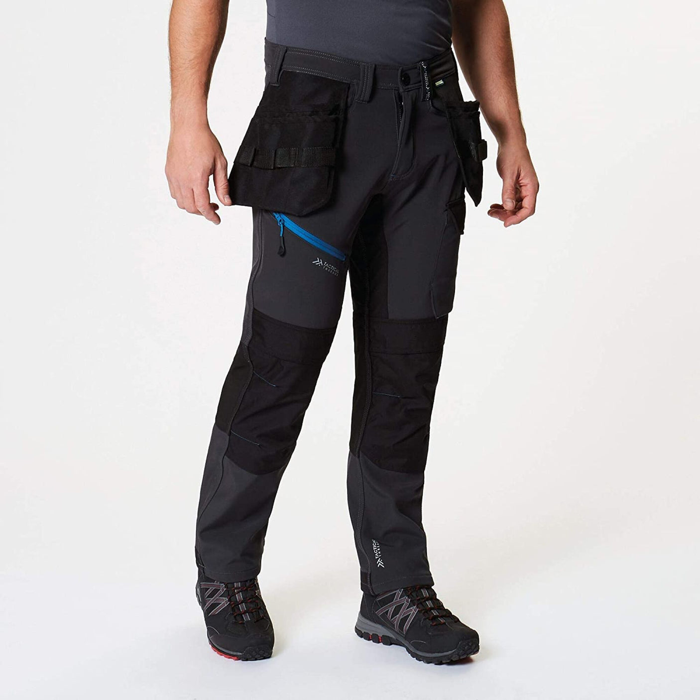 A person wearing Cottonmount's Tactical Threads Strategic Softshell Trouser in Ash Grey, featuring multiple pockets and zippers made from durable CORDURA® fabric, paired with black hiking shoes, stands against a plain background.