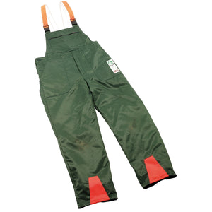 Draper Chainsaw Trousers, Extra Large - CST/N feature green work overalls with orange and white suspenders, bright red patches on the leg bottoms, and an anti-cut inlay that meets EN381 standards against a white background.