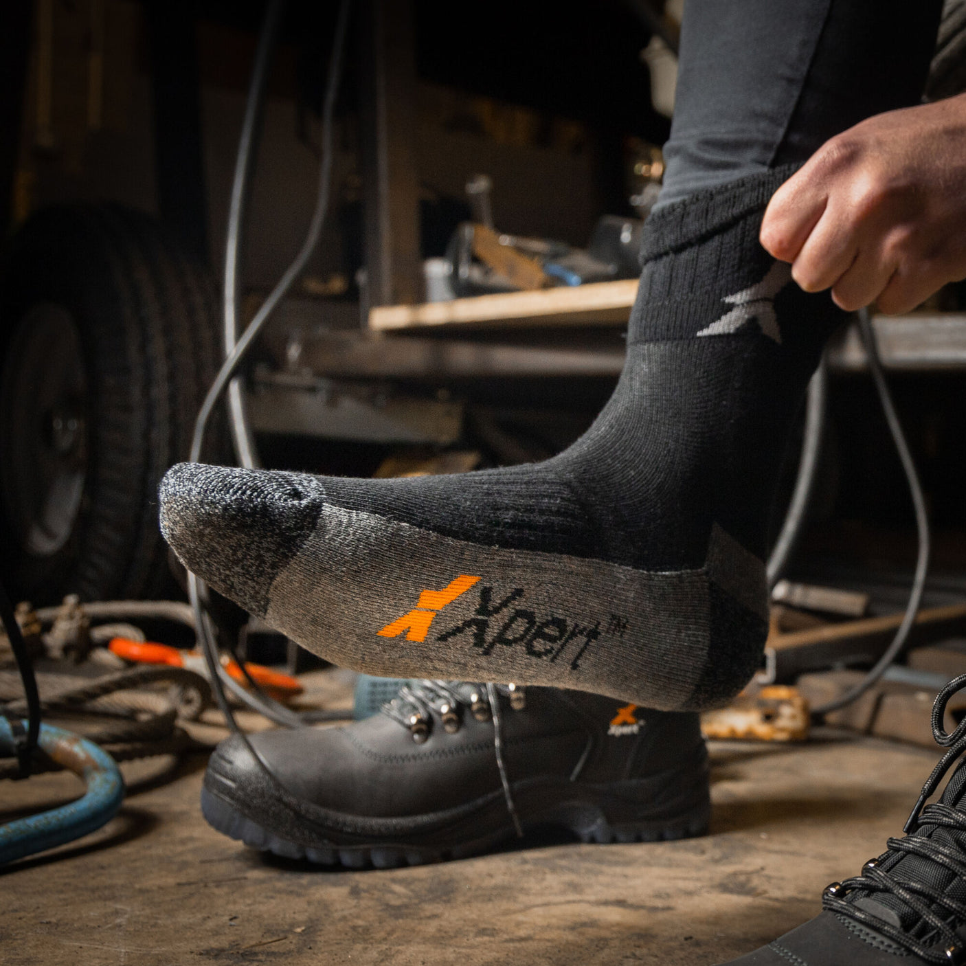 A person is putting on a black Xpert Pro Active Work Sock from the Cottonmount 3 Pack, featuring breathable mesh for maximum comfort, while wearing work boots with the same brand logo in a workshop setting.