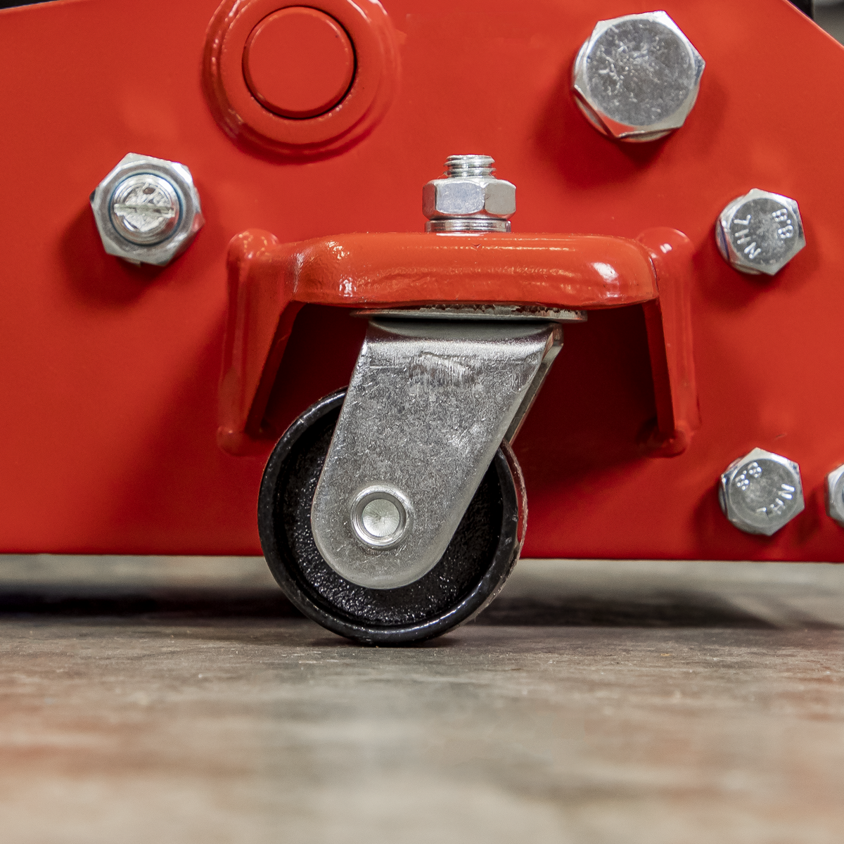 Close-up of a single heavy-duty castor attached to the red metal frame of the Sealey Trolley Jack 3 Tonne Standard Chassis - 3000CXD, with bolts and fasteners visible, resting on a smooth floor surface.