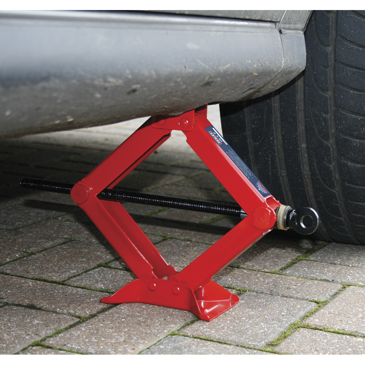 A Sealey Scissor Jack Heavy-Duty 1 Tonne - 57M, featuring a precision thrust bearing, lifts the side of a vehicle near the rear tire on a cobblestone surface, ensuring smooth operation.