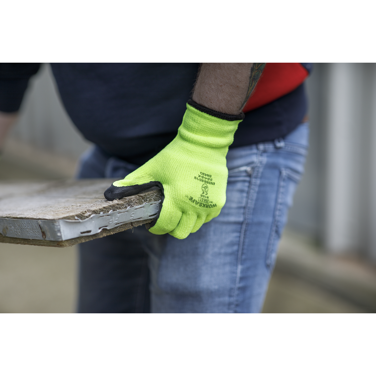 Person wearing Sealey's Thermal Super Grip Gloves (Large) - Pair - 9126 in bright yellow, paired with denim jeans, holding a weathered, rectangular piece of material, possibly a tile, outdoors.