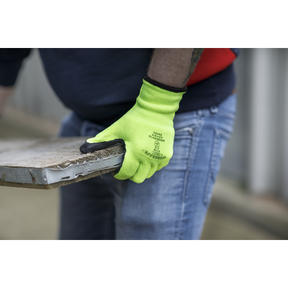 Person wearing Sealey's Thermal Super Grip Gloves (Large) - Pair - 9126 in bright yellow, paired with denim jeans, holding a weathered, rectangular piece of material, possibly a tile, outdoors.