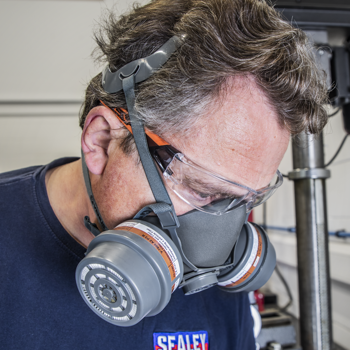 A person wearing safety goggles, a Sealey Half Mask (No Cartridges - 9360) with adjustable straps, and a blue shirt with a "SEALEY" logo is working in a workshop environment.