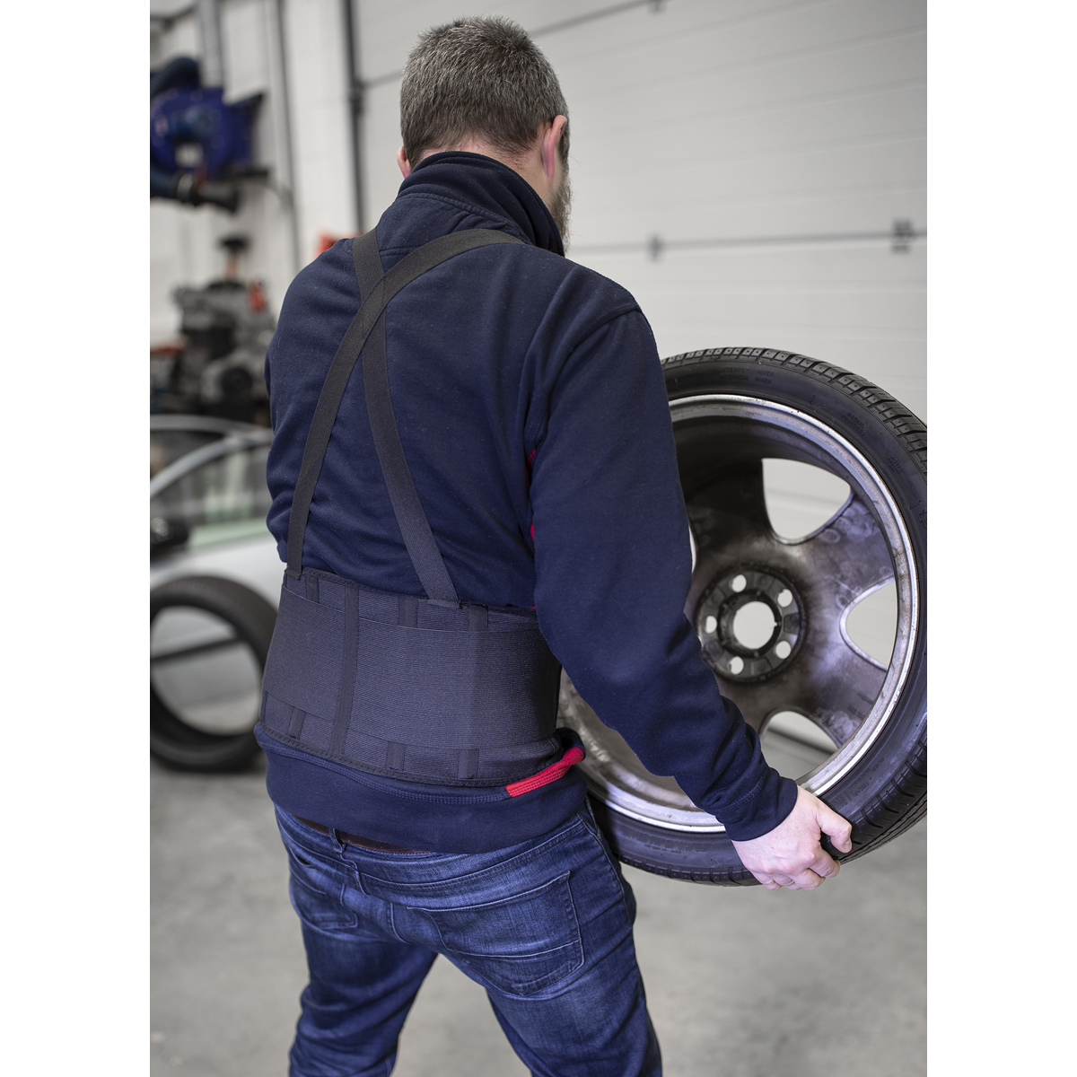 A person in blue jeans is lifting a large car wheel with a tire in a garage, benefiting from all-day support and ease of movement thanks to the Sealey Back Support Belt Large (965-1120mm, model 9707L).