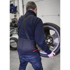 A person in blue jeans is lifting a large car wheel with a tire in a garage, benefiting from all-day support and ease of movement thanks to the Sealey Back Support Belt Large (965-1120mm, model 9707L).