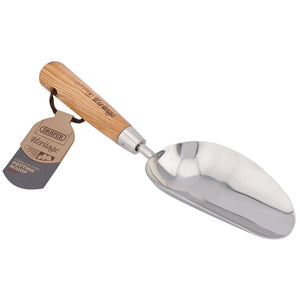 A stainless steel hand potting scoop with an ash handle, crafted in a traditional style and labeled "Draper Heritage Stainless Steel Hand Potting Scoop With Ash Handle - DPSG/L." A branded tag is attached to the handle with a string.