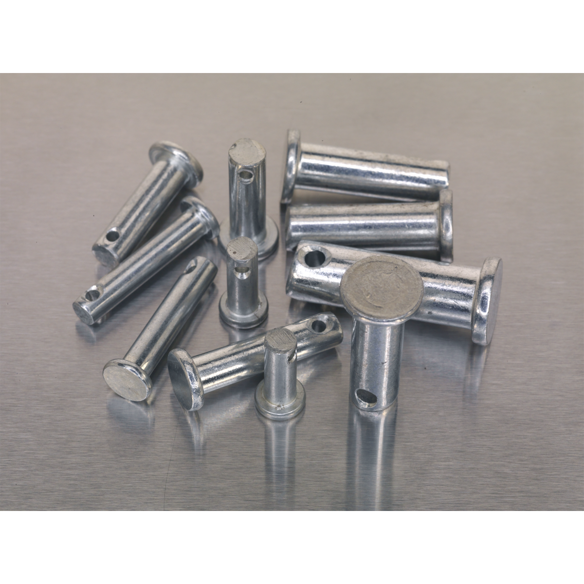 A collection of metallic round-ended cylindrical components with small holes, scattered on a grey surface. This Sealey Clevis Pin Assortment 200pc - Imperial (AB019CP) includes a total of 200 pieces.