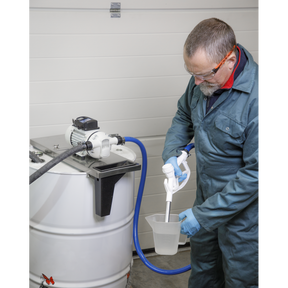A man wearing coveralls, safety goggles, and gloves operates a Sealey Mounting Bracket Drum Pump - ADB09 to fill a measuring container from one of the 205L barrels in an industrial setting.