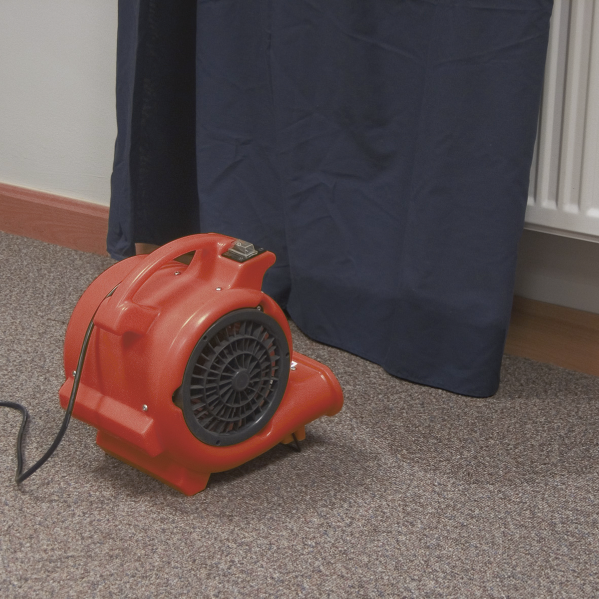 A Sealey Air Dryer/Blower 356cfm 230V - ADB300 with durable composite housing sits on a carpeted floor near a dark curtain and a radiator, ensuring optimal airflow.