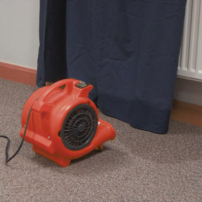 A Sealey Air Dryer/Blower 356cfm 230V - ADB300 with durable composite housing sits on a carpeted floor near a dark curtain and a radiator, ensuring optimal airflow.
