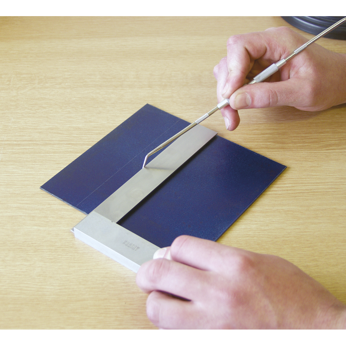 A person uses the Sealey Precision Steel Square 150mm - AK11150 to examine a blue material resting on a flat surface, alongside a metal ruler.