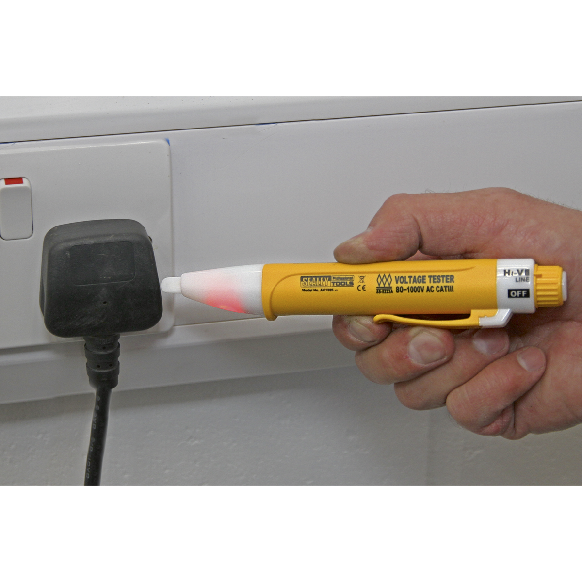 Hand holding a yellow Sealey Non-Contact Voltage Detector 80-1000V - AK1998 with a red light, testing a black electrical plug inserted into a white power outlet. Ideal for detecting wire cable breaks and professional use hand tools.