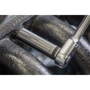 A close-up of a ratchet wrench from Sealey's Socket Set 13pc 1/2"Sq Drive Deep Lock-On™ 6pt Metric - Black Series (AK2745B) being used to tighten a shiny metal bolt on a machine part with a dark, textured surface. The wrench's polished black chrome finish gleams, ensuring both style and durability with its Chrome Vanadium steel construction.