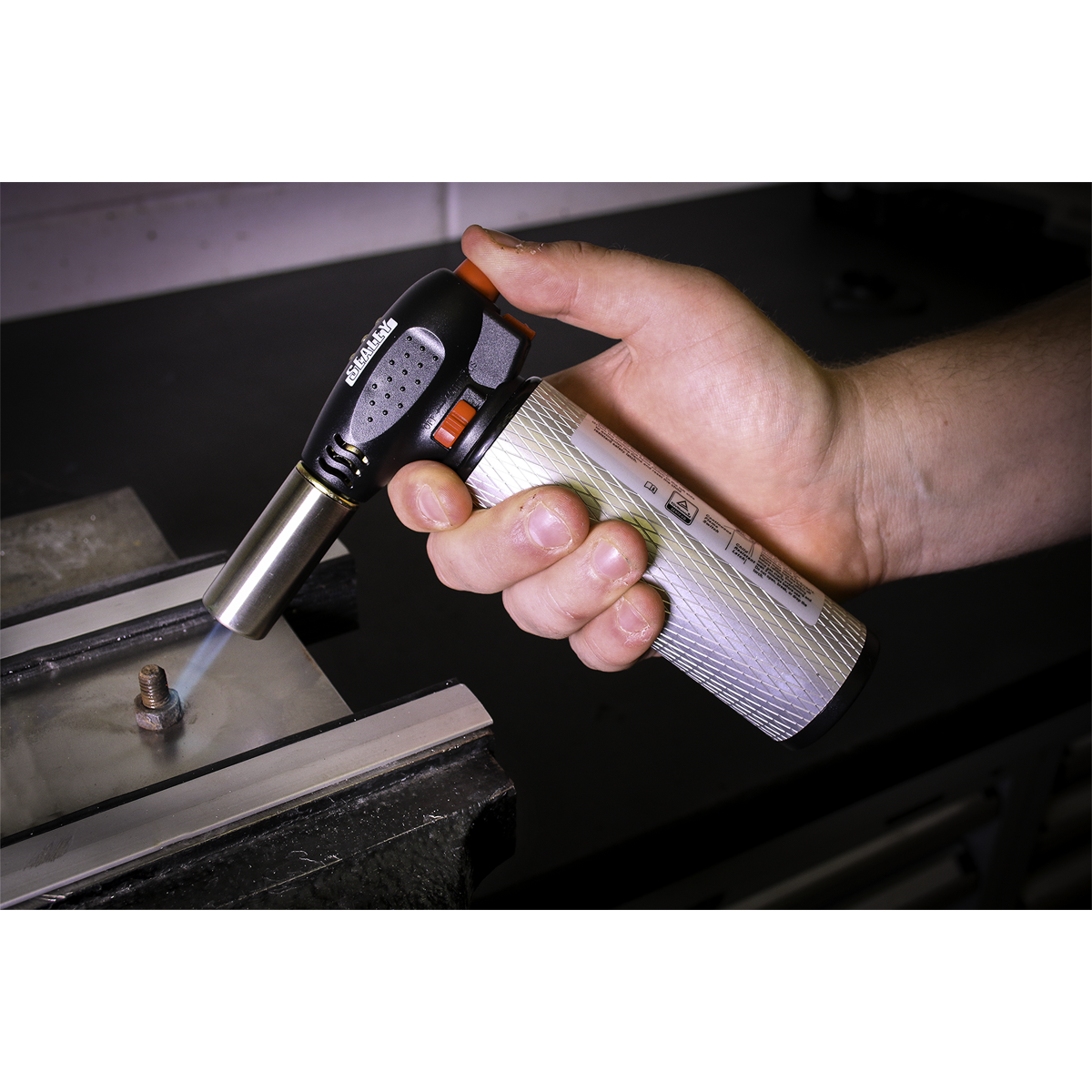 A hand holding the Sealey Heavy-Duty Butane Soldering Torch - AK2932 with piezoelectric ignition, applying heat to a small metal object on a work surface in a workshop.