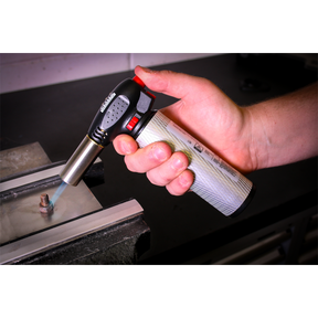 A hand holding the Sealey Heavy-Duty Butane Soldering Torch - AK2932 with piezoelectric ignition, applying heat to a small metal object on a work surface in a workshop.