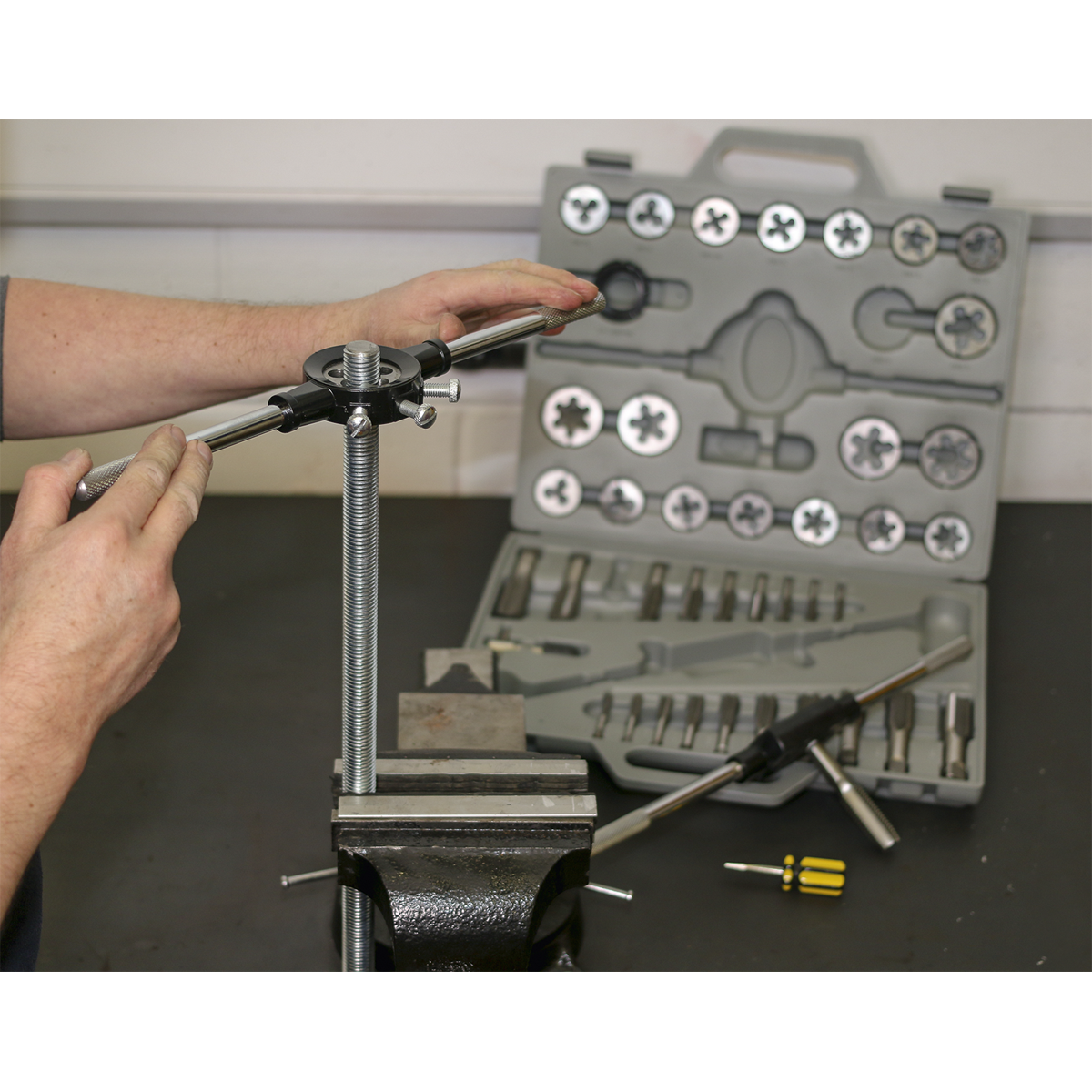 A person is using a threading tool on a metal rod, held in a vise. A Sealey Tap & Die Set 45pc Split Dies Metric - AK303, featuring various Metric sizes and Tungsten steel taps and dies, is open on the table in the background.